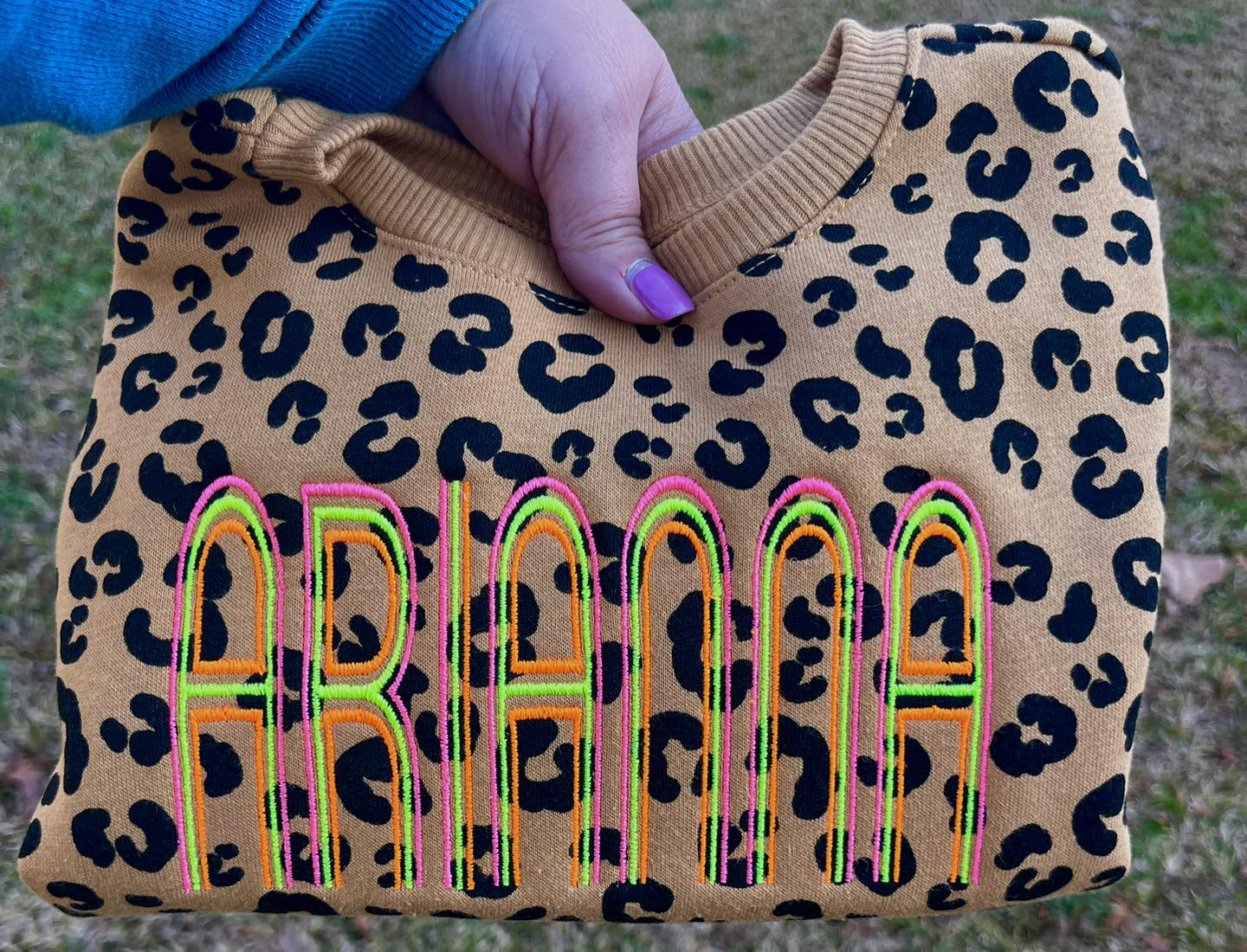 Leopard print sweatshirt with colorful neon pink, yellow and orange embroidered name Arianna.  Sweatshirt is being held by a person for display