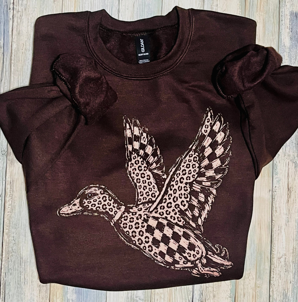 leopard and check design on a flying mallard duck decal.  decal is light brown and pressed onto a dark brown sweatshirt  sweatshirt is lying on a wooden background