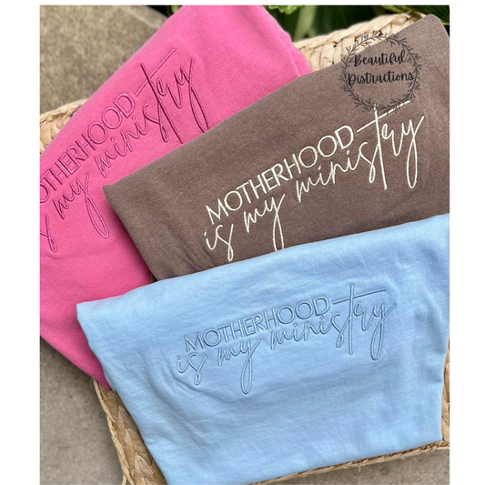pink, brown and blue tees with motherhood is my ministry embroider on them in coordinating thread  lying on a whicker pad  