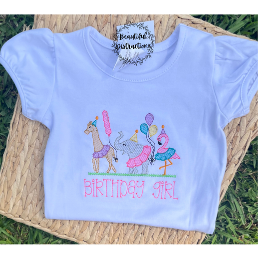 Embroidered design of a giraffe, elephant, and flamingo with balloons and 'Birthday Girl' text on a woven background lying in the grass