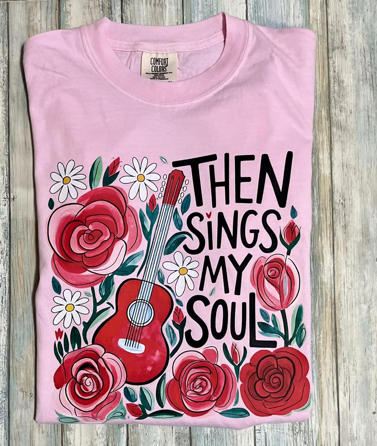 Pink t-shirt with floral design and text 'Then Sings My Soul' on a wooden surface