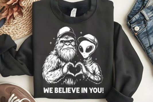 Black sweatshirt with a graphic of a bigfoot and alien holding hands and text 'WE BELIEVE IN YOU!' on a white surface.