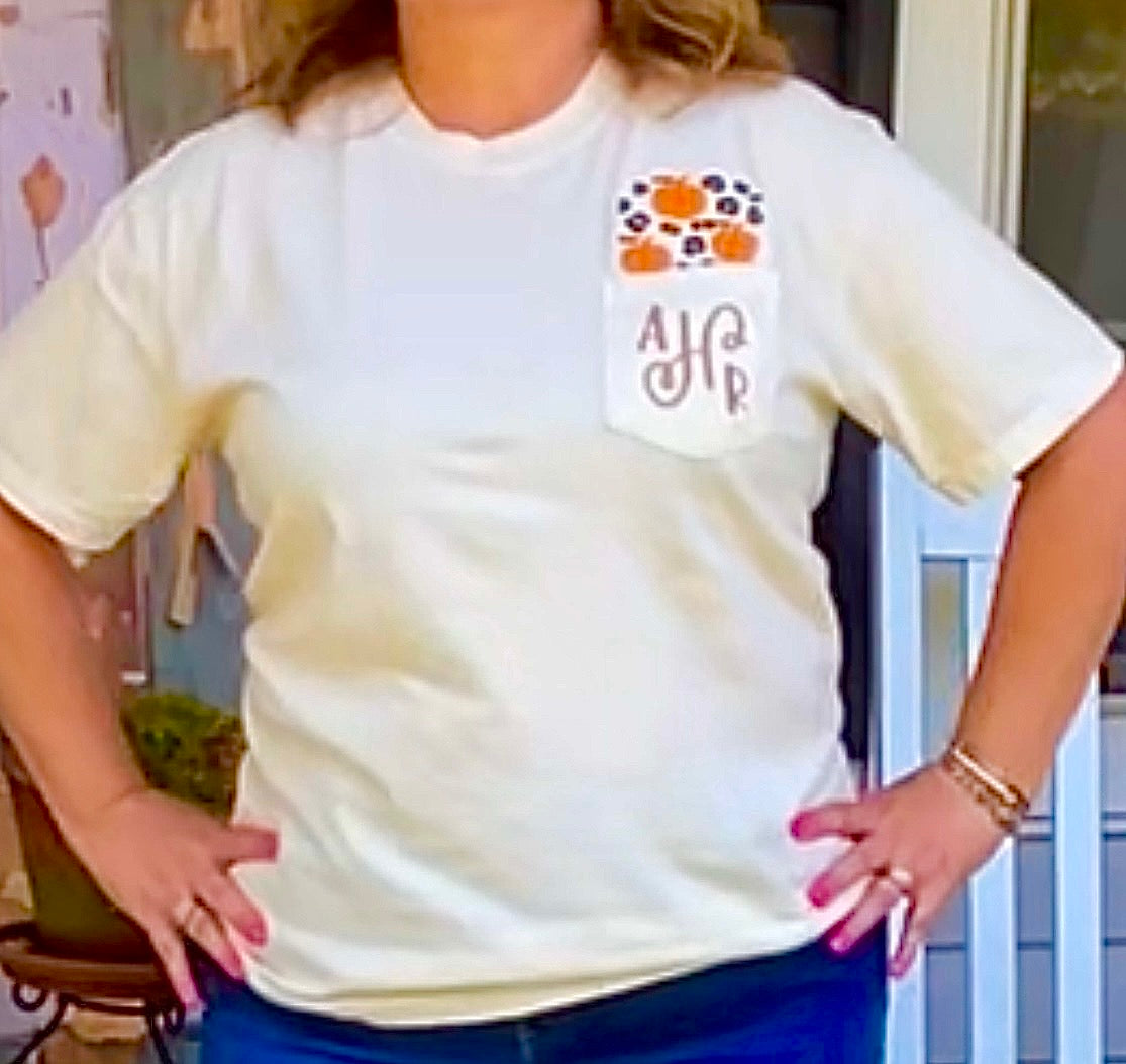 woman with hands on hips wearing ivory short sleeve tee     tee has pumpkins and leopard spots embroidered above the pocket and the initials AHR embroidered on the pocket
she is standing in front of a door